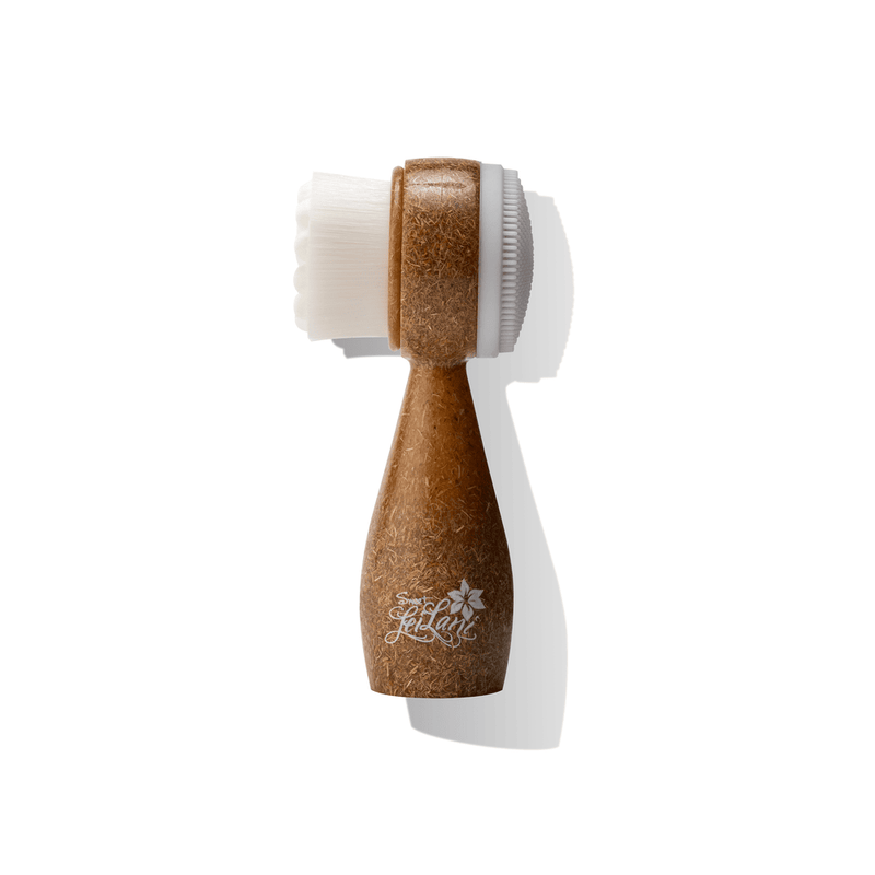 A dual-sided facial cleansing brush with soft bristles on one side and a silicone exfoliating pad on the other, featuring a wooden handle and white bristles.