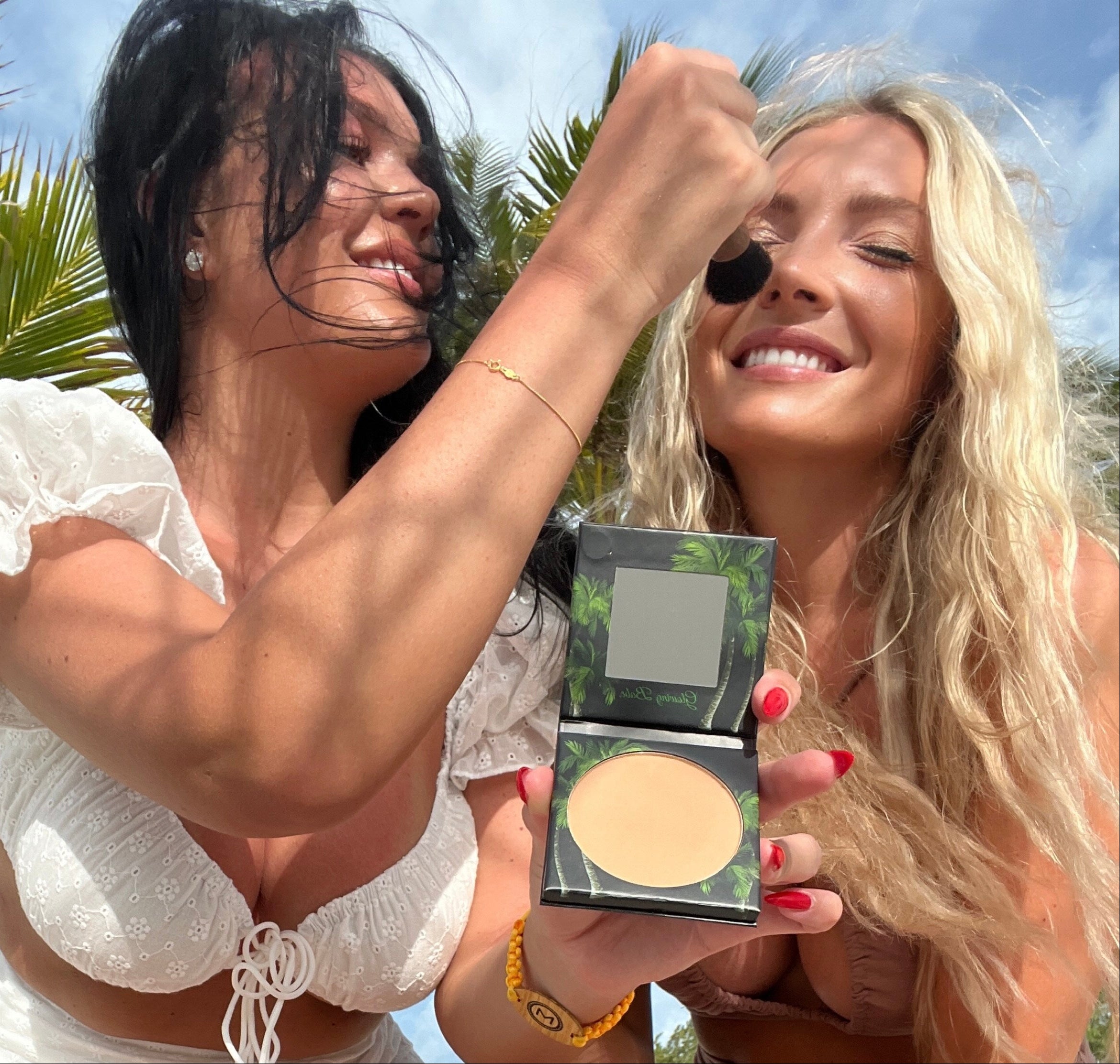 Two women outdoors with palm trees, one applying makeup to the other.