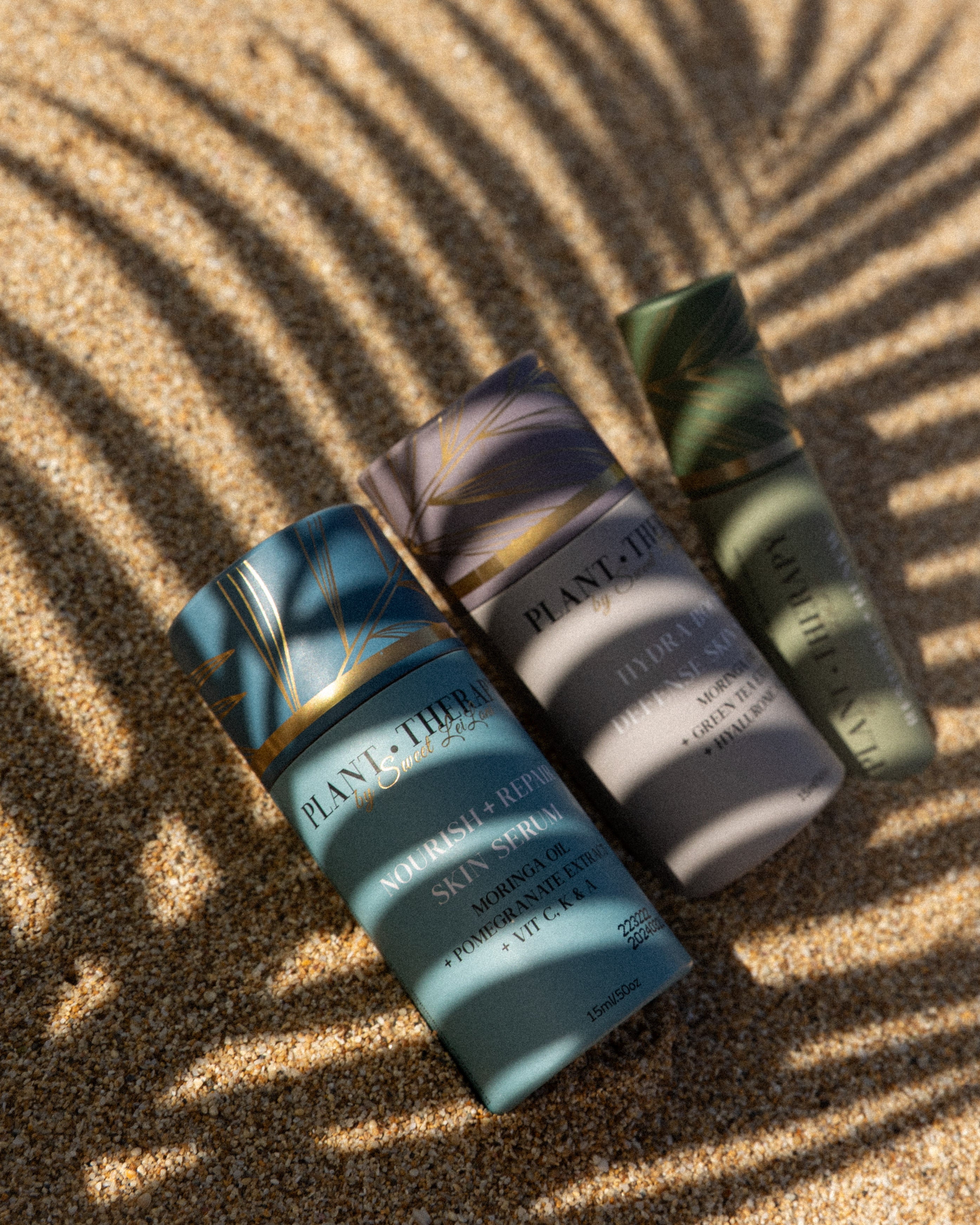 A collection of Plant Therapy skincare products including serums and a roll-on elixir, displayed on a sandy surface with a shadow of a plant leaf across them.